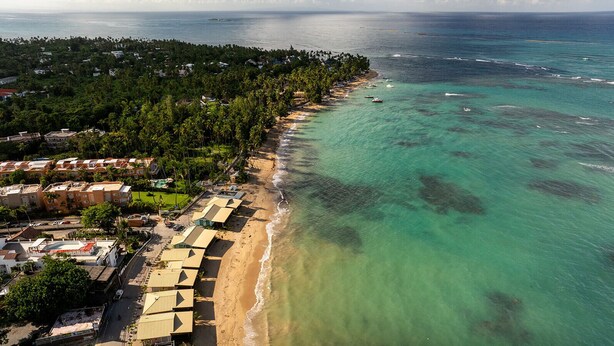 An aerial view of a coastal area with a beachfront hotel, clear turquoise waters, and a dense forested area in the background.