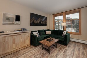A living room with a green sectional sofa, a wooden coffee table, a mounted TV, and a large window with a view of buildings and trees. - NEW! Premium Unit 2216 - 1 Br - Zephyr Mountain Lodge (Winter Park)
