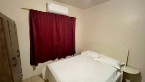 Ein Schlafzimmer mit einem Bett, weißen Vorhängen, einem weißen Kopfende, einem Nachttisch und einer Klimaanlage.