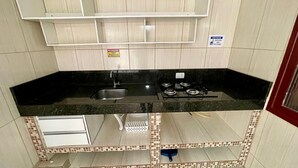 Une cuisine avec un plan de travail noir, un évier en acier inoxydable et une cuisinière à gaz. On trouve un petit placard sous le plan de travail et un pare-feu en carrelage.