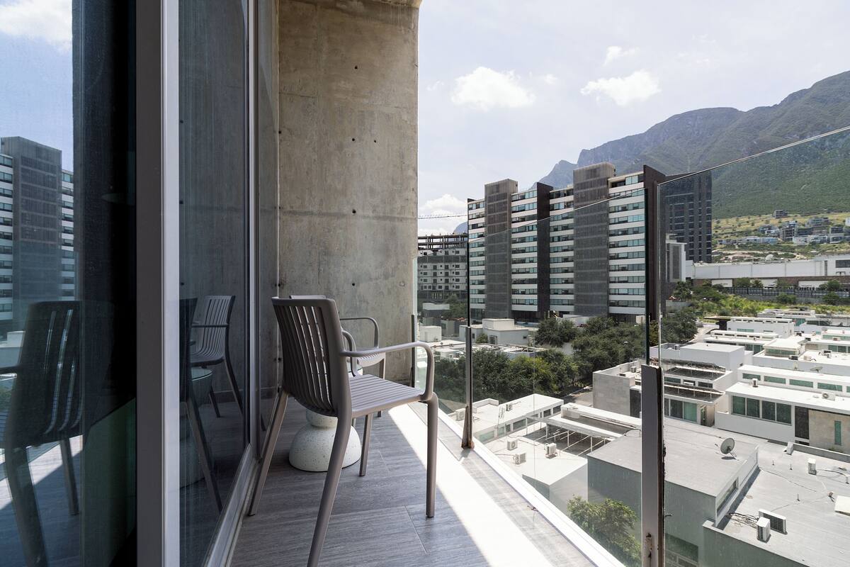 Un balcón con dos sillas, un pilar de hormigón y vistas a edificios modernos y montañas.