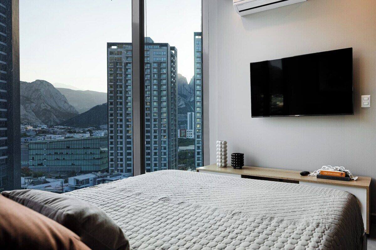 Un dormitorio con una cama grande, un televisor de pantalla plana montado en la pared y vistas a un paisaje urbano con edificios altos.