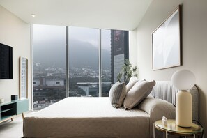 A modern bedroom with a large bed, a bedside table, a wall-mounted TV, and a large window offering a city view. - Modern Apts w Pool & Mountain View (Santa Catarina)