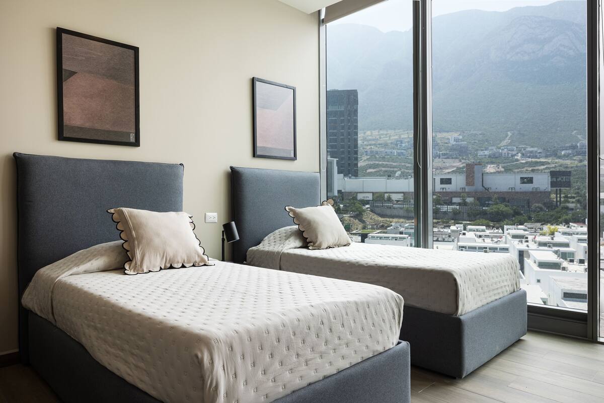 Habitación de hotel con dos camas, un ventanal amplio que ofrece vistas a la ciudad y a la montaña, y cuadros enmarcados en la pared.