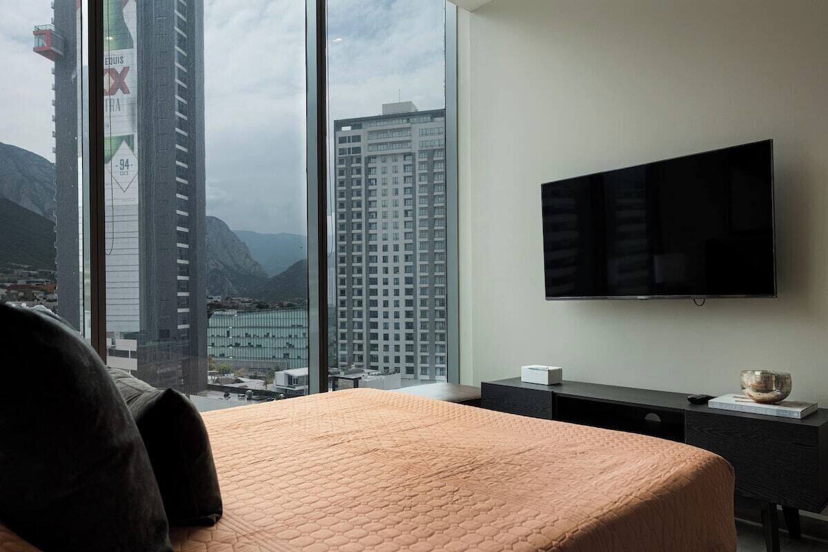 Habitación de hotel con una cama grande, un televisor de pantalla plana en la pared y vistas a edificios de la ciudad y montañas a través de la ventana.