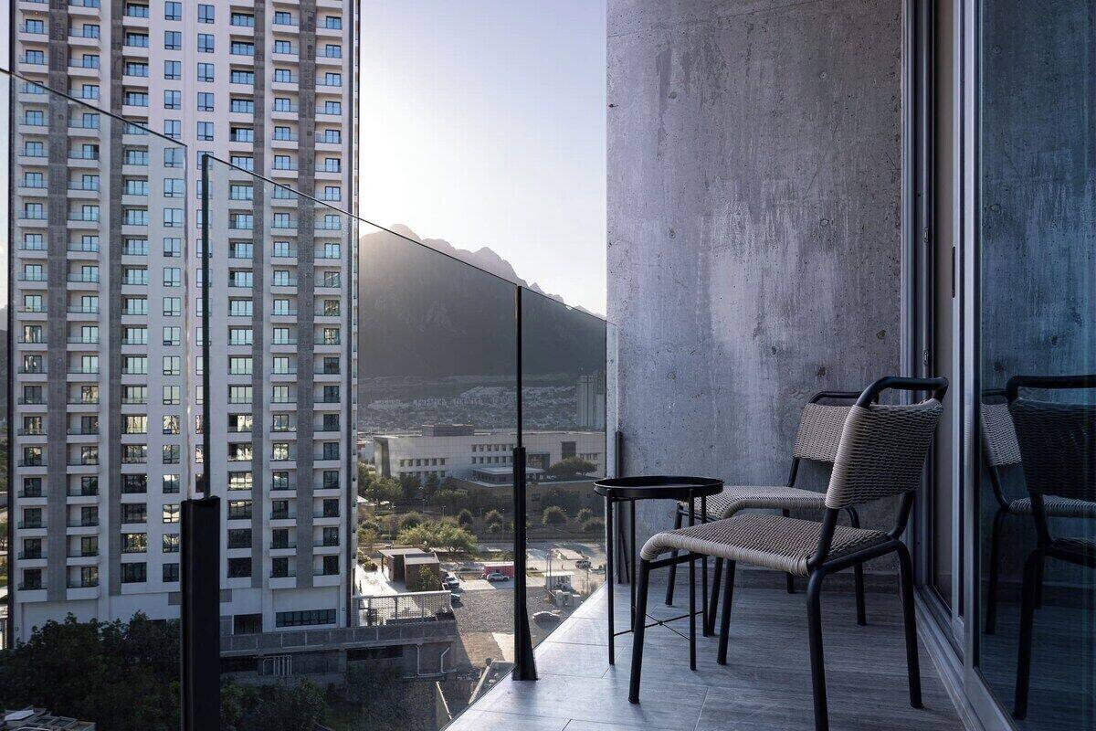 Un balcón con dos sillas de mimbre y una pequeña mesa, con vistas a un paisaje urbano con edificios altos y una montaña distante.