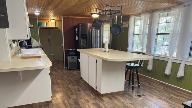 A kitchen with a wooden floor, white cabinets, a stainless steel refrigerator, a water dispenser, and a hanging pot rack.