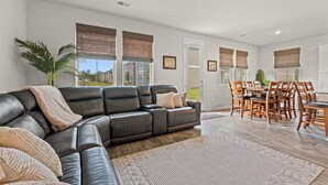 A modern living room with a leather sectional sofa, dining area with wooden chairs, large windows with blinds, and a ceiling fan.