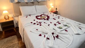 A neatly made bed with rose petals and towels arranged in a heart shape, a bedside lamp, and a bottle of wine on the nightstand.