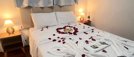 A neatly made bed with rose petals and towels arranged in a heart shape, a bedside lamp, and a bottle of wine on the nightstand.