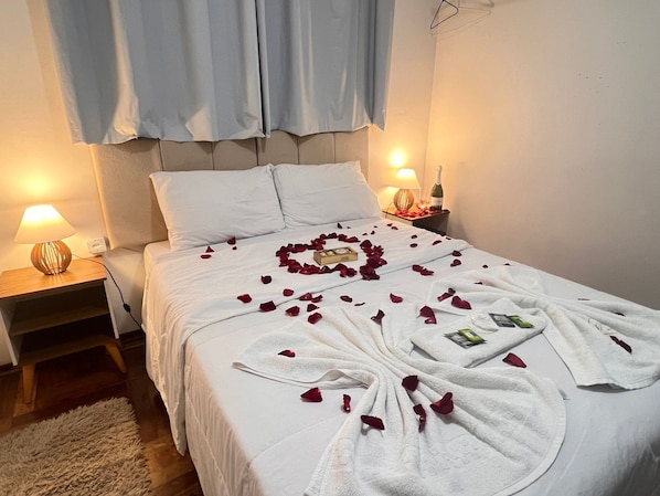A neatly made bed with rose petals and towels arranged in a heart shape, a bedside lamp, and a bottle of wine on the nightstand.