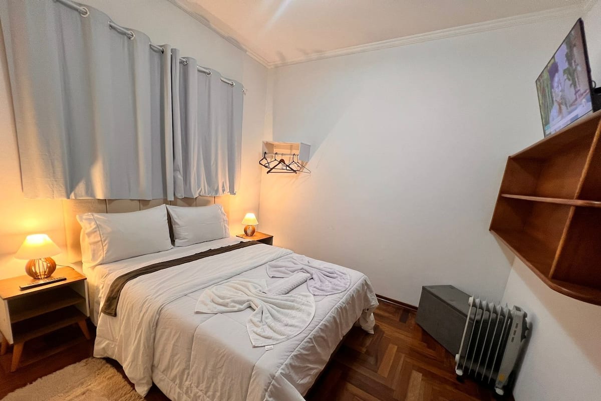 Um quarto arrumado com cama, abajures, radiador e prateleira na parede.