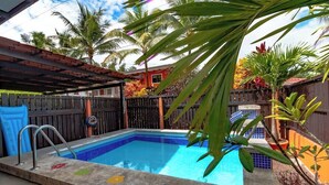 Une piscine dont le bord est recouvert de carrelage et qui est équipée d’un escalier en métal, le tout entouré d’une clôture en bois et de palmiers.