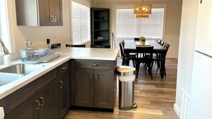 A modern kitchen with dark cabinets, a stainless steel sink, and a dish rack. There is a dining area with a table and chairs, and a trash can.
