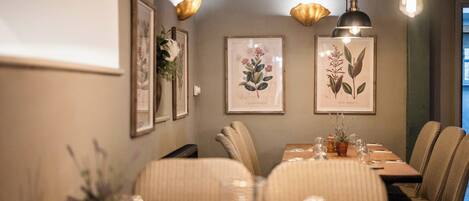 A dining area with a long table set for six, surrounded by upholstered chairs. There are framed botanical prints on the wall, and pendant lighting hangs above.