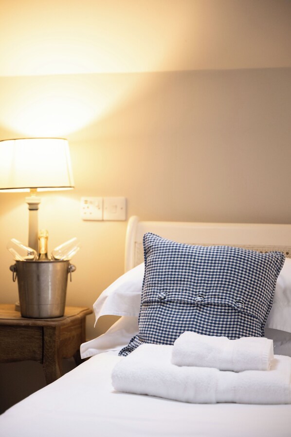 A neatly made bed with white linens and a checkered pillow, a bedside lamp, and a metal container with a bottle. - St Tudy Inn (Bodmin)