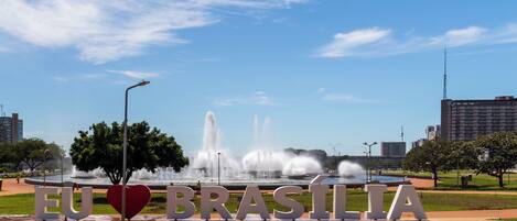 Un parque con una fuente, letras grandes que dicen 'EU BRASÍLIA' con un símbolo de corazón, y un cielo azul despejado.