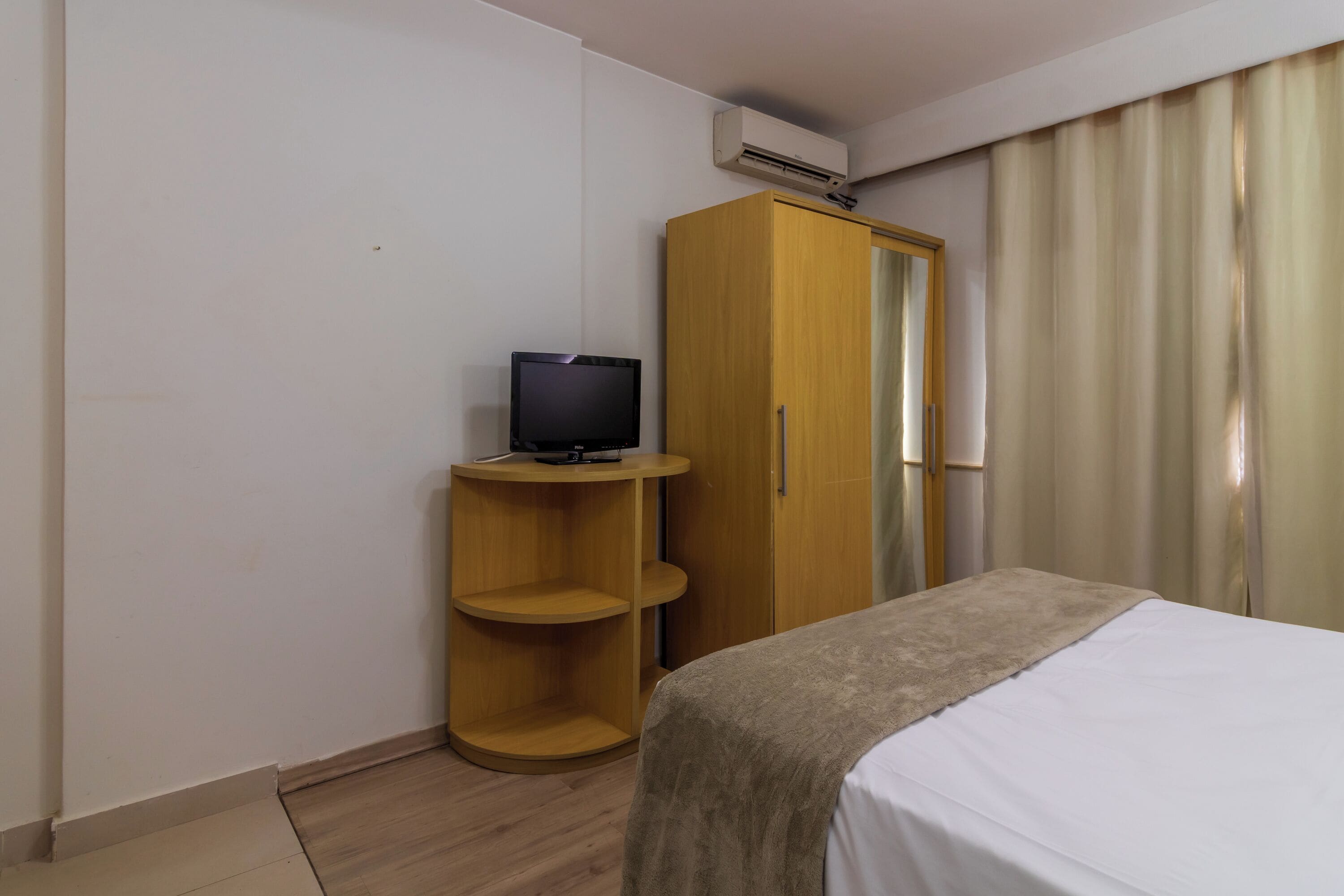 A hotel room with a bed, a wooden wardrobe, a television on a stand, and an air conditioning unit on the wall.