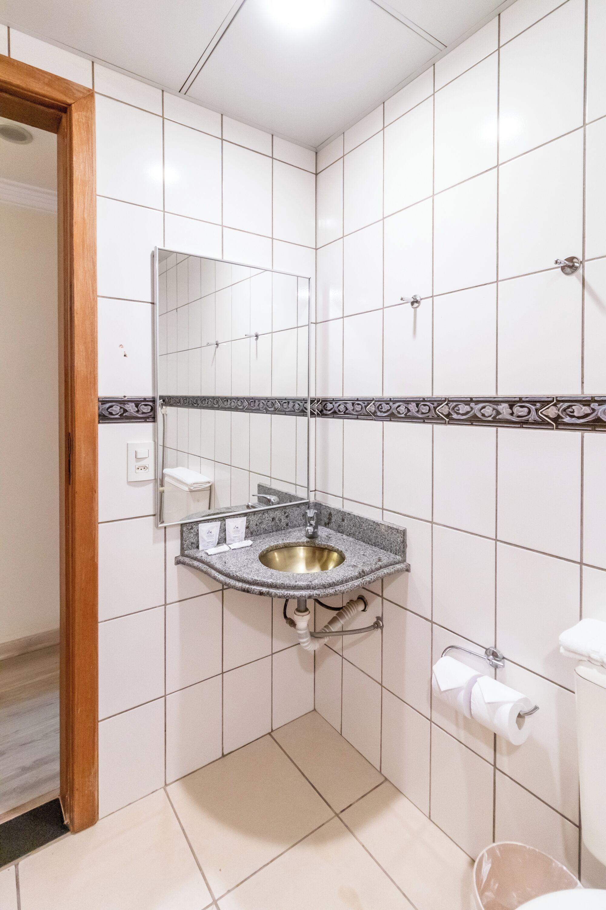 Un baño con paredes de azulejos blancos, un lavabo de granito, un espejo, un perchero para toallas y un inodoro.