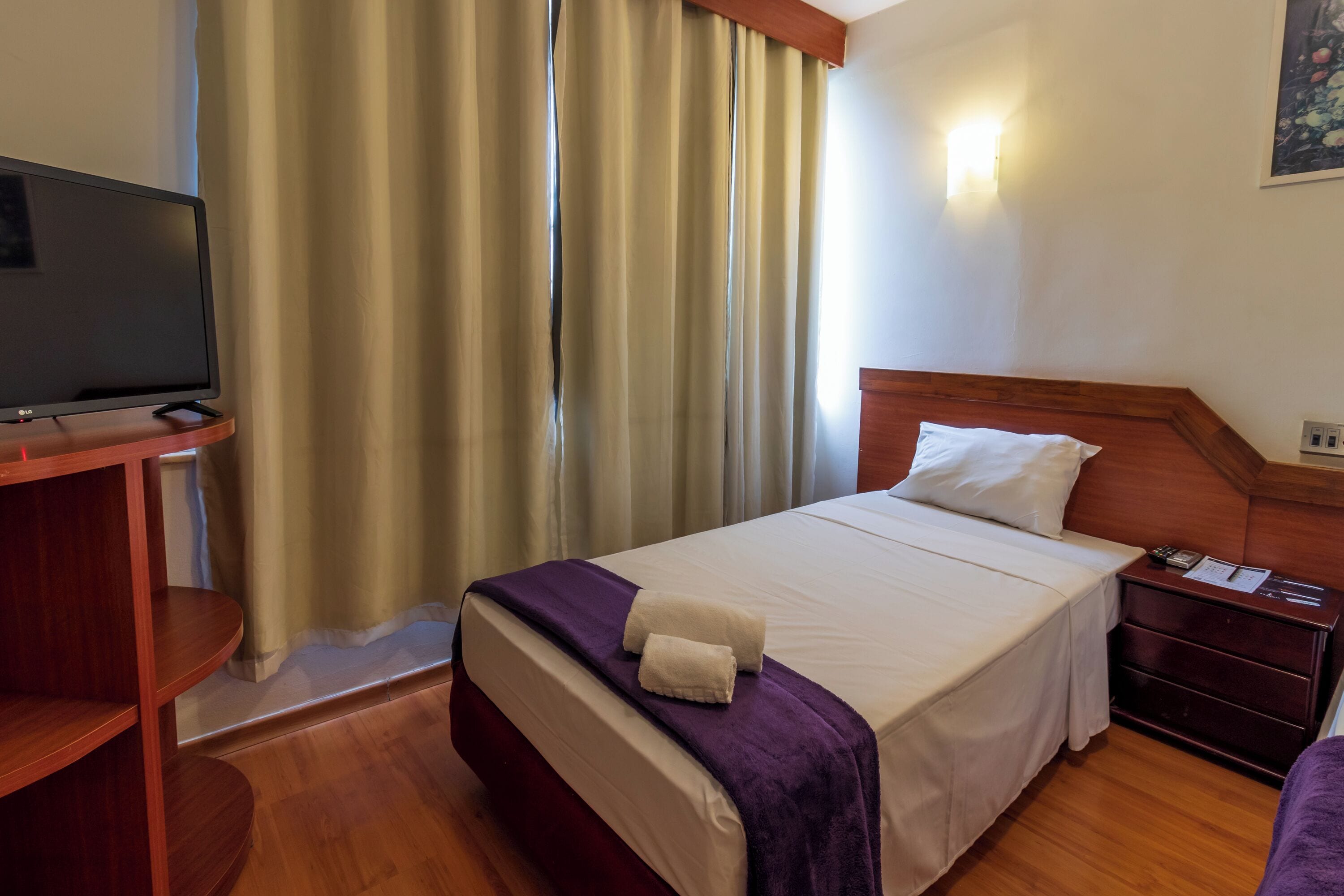 A hotel room with a bed, a TV on a wooden stand, a nightstand with a lamp, and a window with curtains.