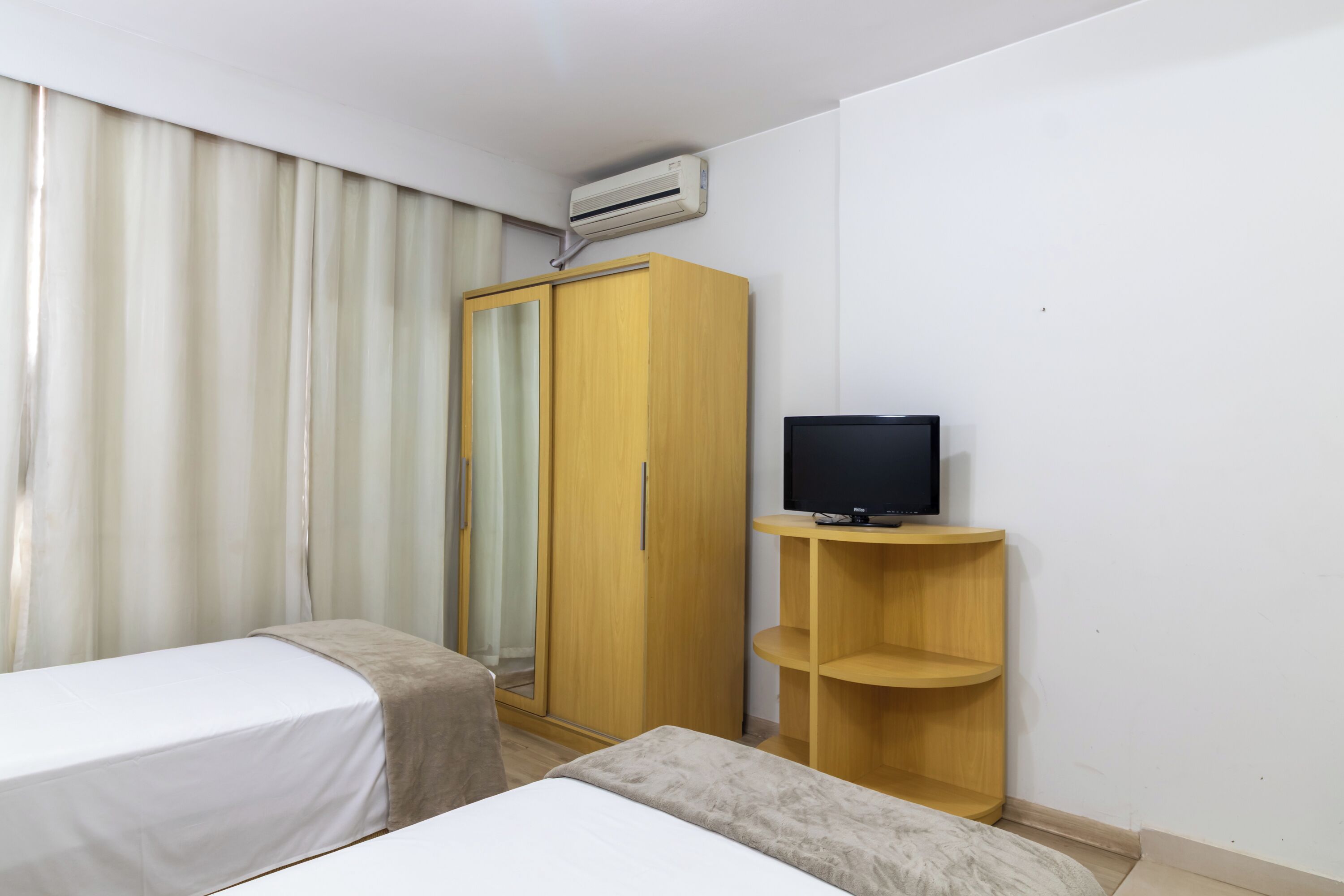Een hotelkamer met twee enkele bedden, een garderobe, een televisie op een houten standaard en een airconditioner.