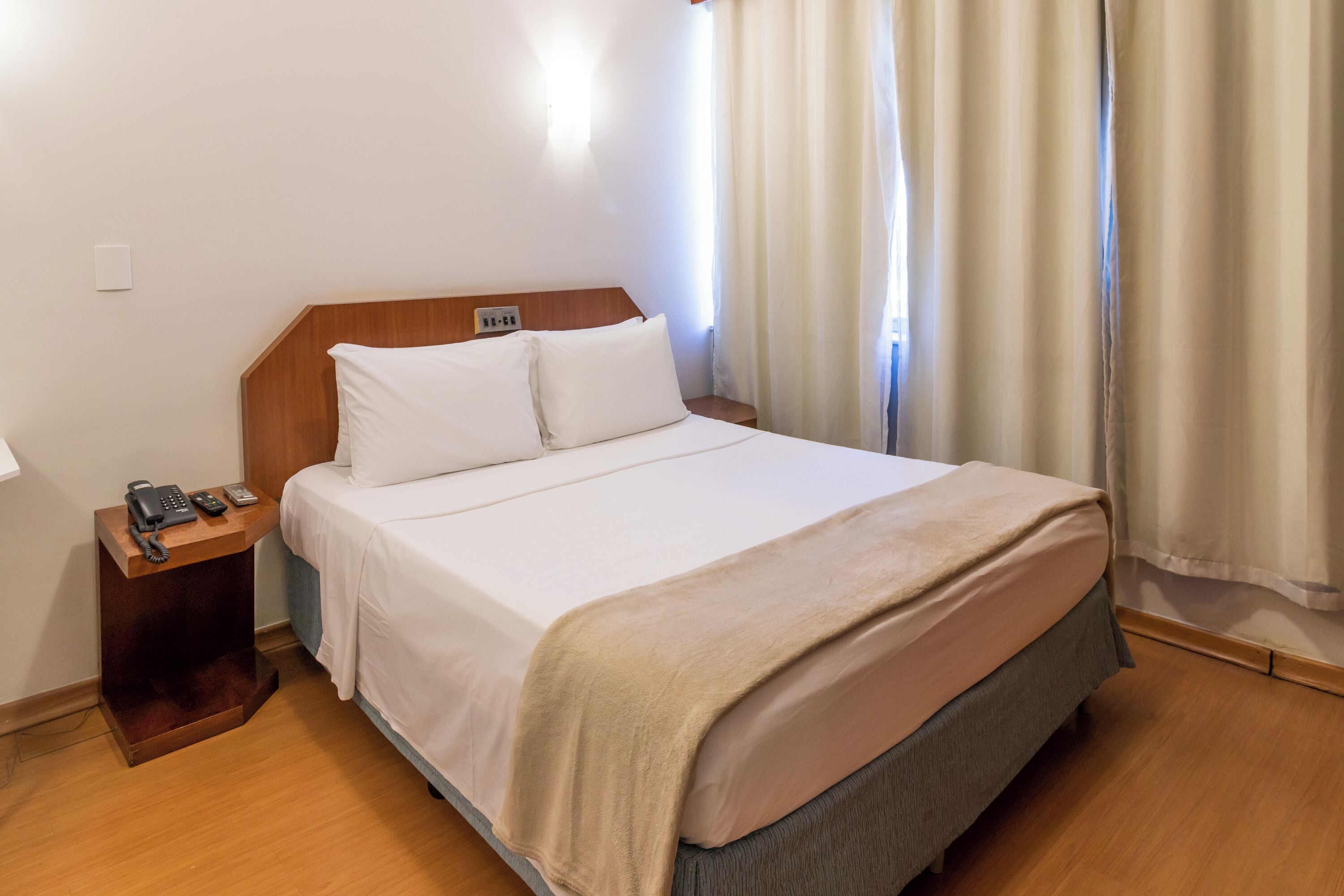 Quarto de hotel com cabeceira de madeira, roupa de cama branca, mesa de cabeceira com telefone e controle remoto, e janela com cortinas.