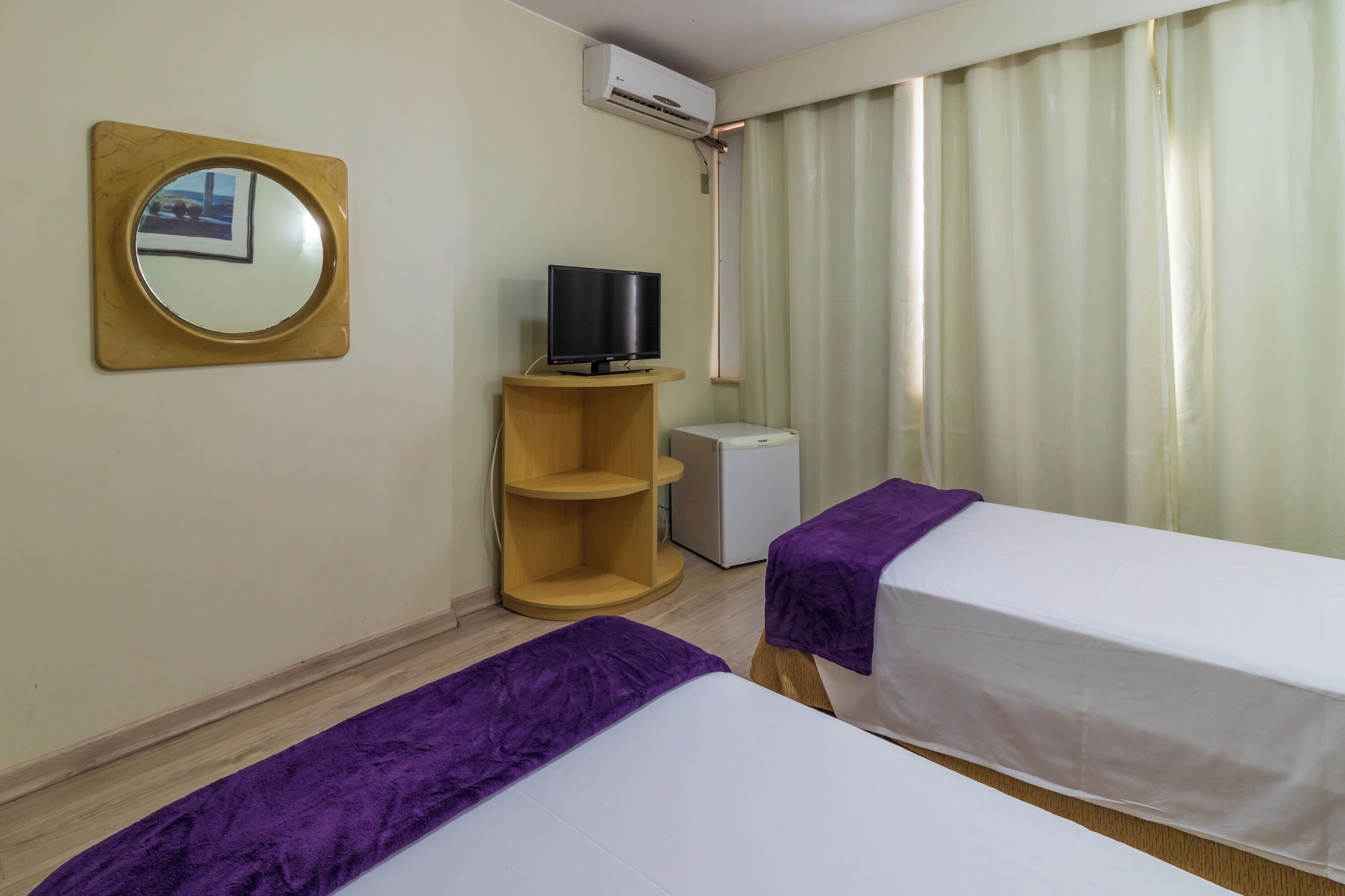 Une chambre d’hôtel avec deux lits, une télévision, une petite étagère en bois, un appareil de climatisation et une fenêtre avec des rideaux.