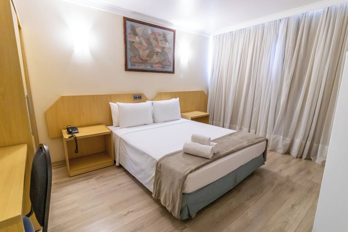 Ein Hotelzimmer mit einem Bett, einem Nachttisch, einem Sessel, einem Gemälde an der Wand und einem Fenster mit Vorhängen.
