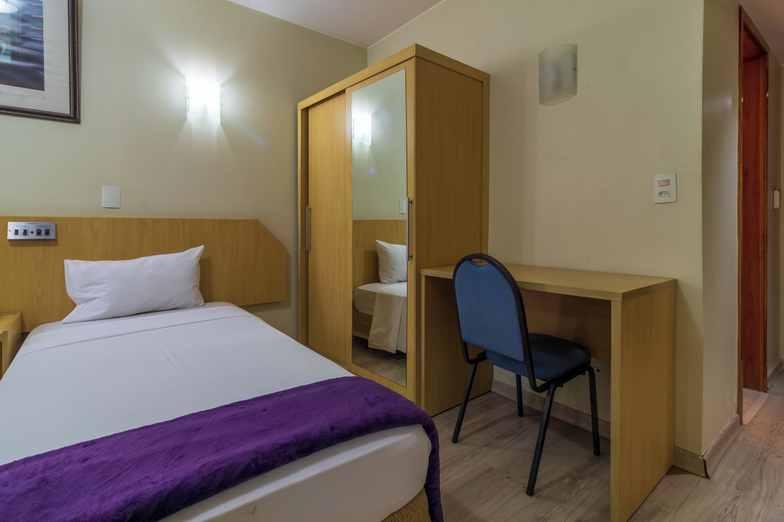 A hotel room with a bed, a desk, a chair, a wardrobe, and a bedside table.