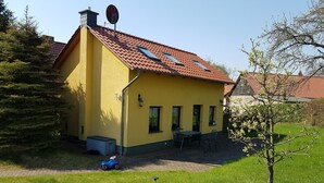 Una casa amarilla con techo de tejas rojas, una antena parabólica y un pequeño patio con mesa y sillas.