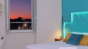 A modern hotel room with a city view at dusk, a neatly made bed, and a teal accent wall. - Domus Augustea San Giovanni Sunset Suites - Deluxe Suite Ottanivm (Roma)