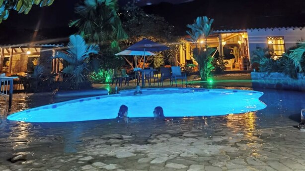 Zona de piscina nocturna con dos personas en el agua, rodeada de palmeras e un edificio con iluminación cálida.