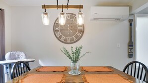 A dining area with a wooden table, orange placemats, a large wall clock, hanging pendant lights, and a vase with greenery.