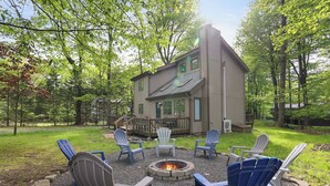 A house with a fire pit surrounded by Adirondack chairs in a wooded area.