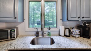 A kitchen with a stainless steel sink, a microwave, a coffee maker, and a window with a view of trees.