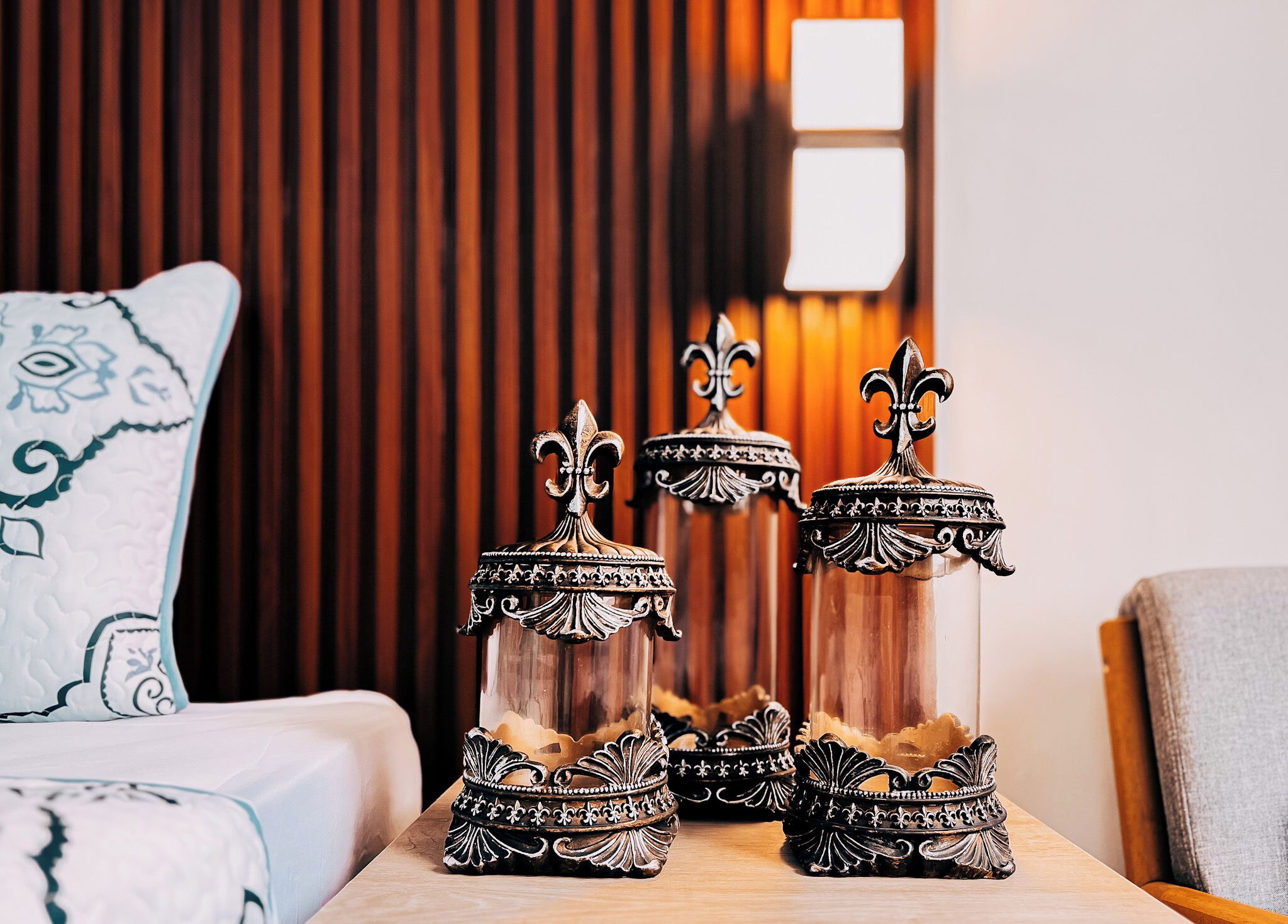 A table with three ornate candle holders with fleur-de-lis designs, placed on a wooden table in a room with a brown curtain and a white pillow with a blue pattern.