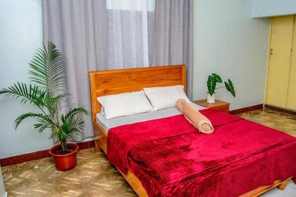A neatly made bed with a red blanket, a wooden headboard, and two white pillows. There is a potted plant to the left of the bed and another on the right side of the room.