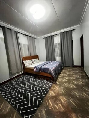 A bedroom with a bed, grey curtains, a patterned rug, and a wooden floor. - Bustani Luxury Homestay (Moshi)