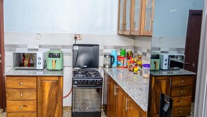 A compact kitchen with a gas stove, two microwaves, a kettle, and various kitchen items on the counter. - Bustani Luxury Homestay (Moshi)