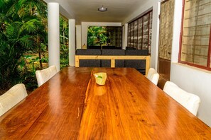 A wooden dining table with white cushioned chairs, a woven basket, and a view of a lush garden through a glass partition. - Bustani Luxury Homestay (Moshi)