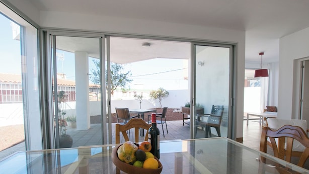 Un salon moderne avec une table en verre, un saladier rempli de fruits et une porte-fenêtre coulissante donnant sur une terrasse extérieure aménagée avec du mobilier.