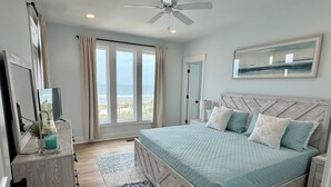 A bedroom with a bed, a ceiling fan, a nightstand, a lamp, a dresser, a window with curtains, and a painting on the wall. - NewBeach Front Home,6bd,6.5ba Elevator,Pool,Spa!! (Ocean Isle Beach)