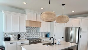 A modern kitchen with white cabinetry, a central island with stools, a stainless steel refrigerator, and a built-in oven. - NewBeach Front Home,6bd,6.5ba Elevator,Pool,Spa!! (Ocean Isle Beach)