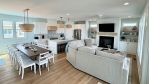 A spacious living area with a large sofa, a dining table with chairs, a kitchen with white cabinets, and a fireplace. - NewBeach Front Home,6bd,6.5ba Elevator,Pool,Spa!! (Ocean Isle Beach)