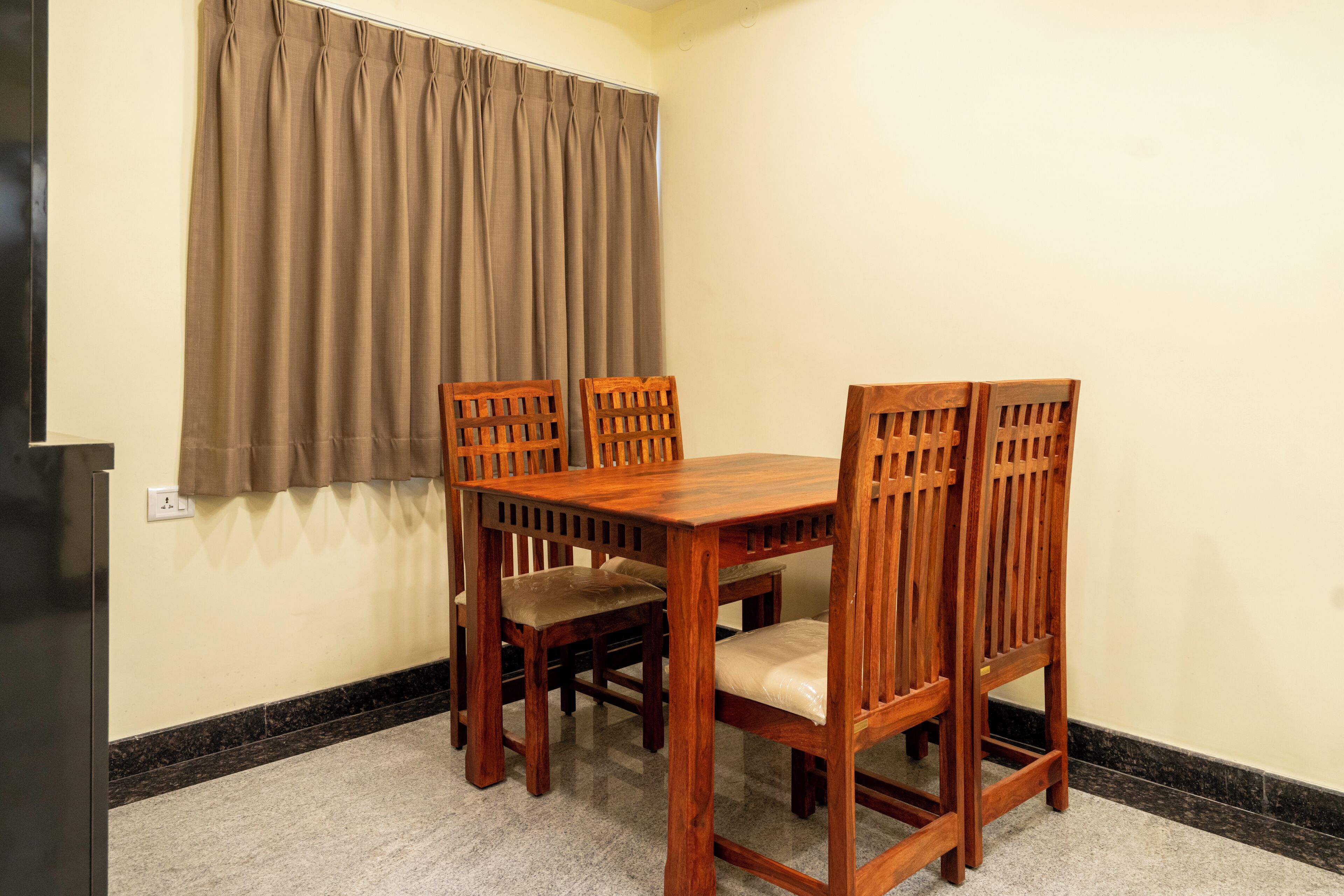 A dining area with a wooden table and four chairs, a brown curtain, and a black baseboard.