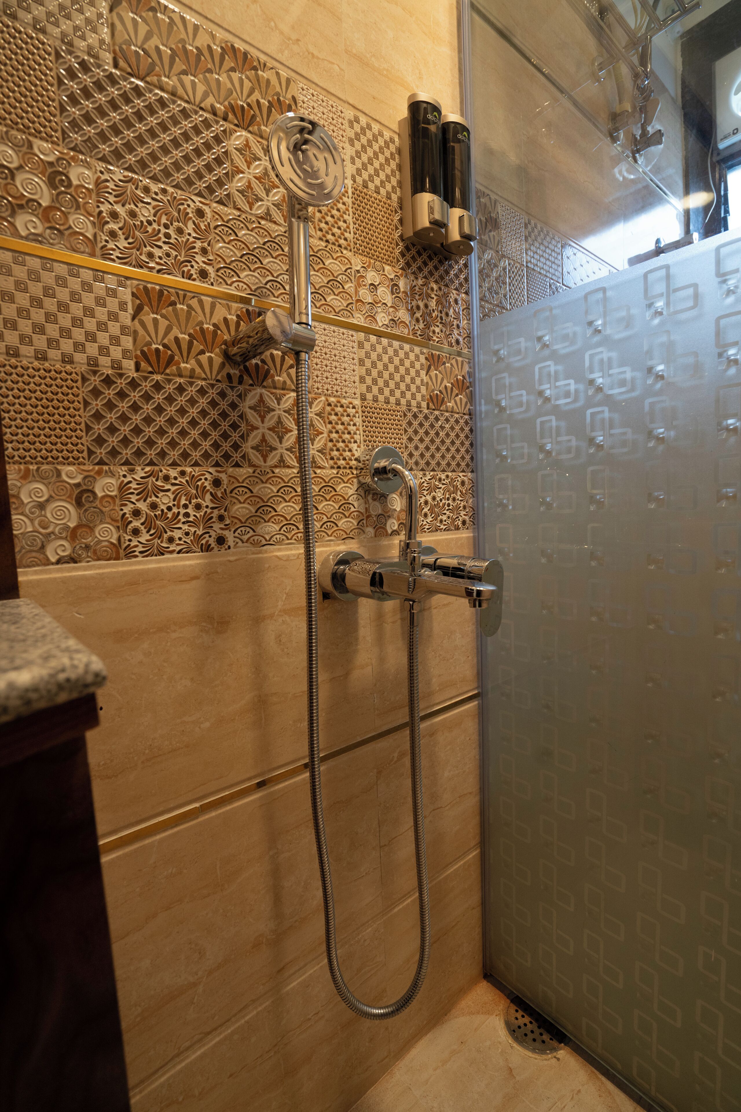 Une douche équipée d’un pommeau de douche à main et d’un robinet mural. L’espace douche est agrémenté de carreaux décoratifs sur les murs et d’une porte en verre.