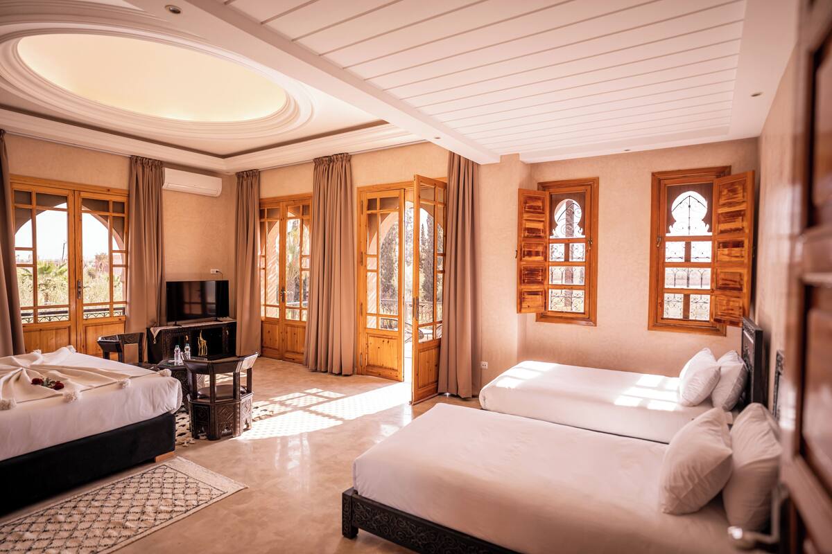 A hotel room with two beds, a TV, a desk, and large windows with curtains.