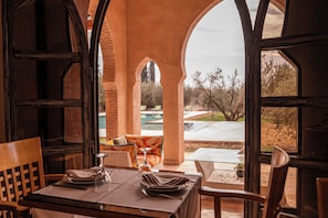 Comedor con mesa lista para dos, con vista a una piscina y zona de estar al aire libre.