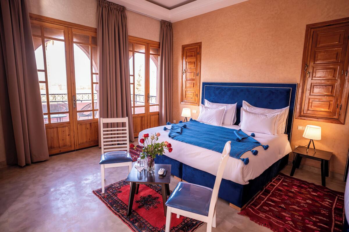 A hotel room with a large bed, a chair, a small table with a vase of flowers, and a view through the window.
