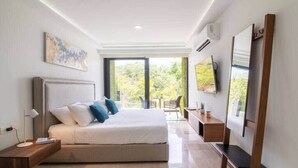 Een moderne slaapkamer met een groot bed, een bureau, een televisie en een balkon met uitzicht op groen.